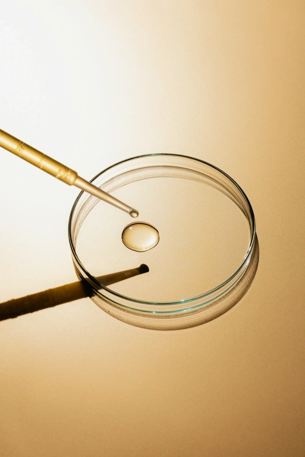 A golden dropper dispenses a clear liquid droplet into a glass petri dish, set against a warm, reflective golden background.