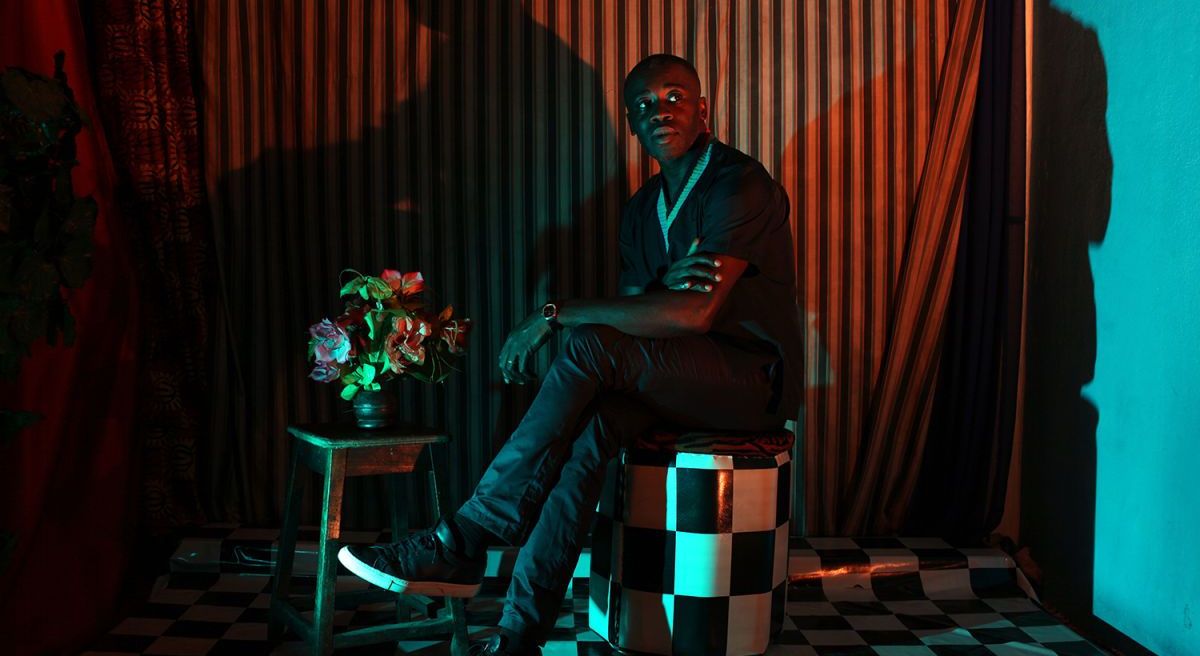 Man seated on a checkered stool in a dramatically lit room with striped curtains, casting bold shadows, next to a small table holding a vase of flowers