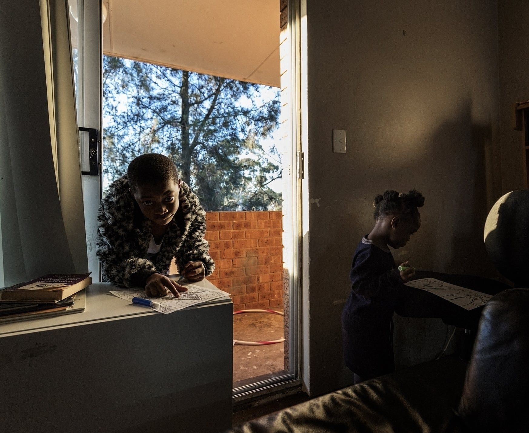 Two children doing homework indoors, one leaning by a window and the other drawing at a desk, with warm sunlight casting shadows.