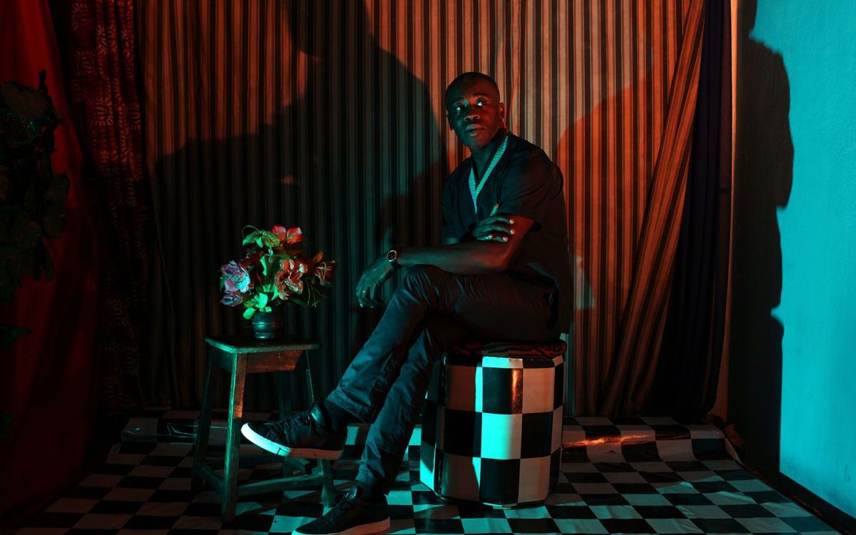 Man seated on a checkered stool in a dramatically lit room with striped curtains, casting bold shadows, next to a small table holding a vase of flowers