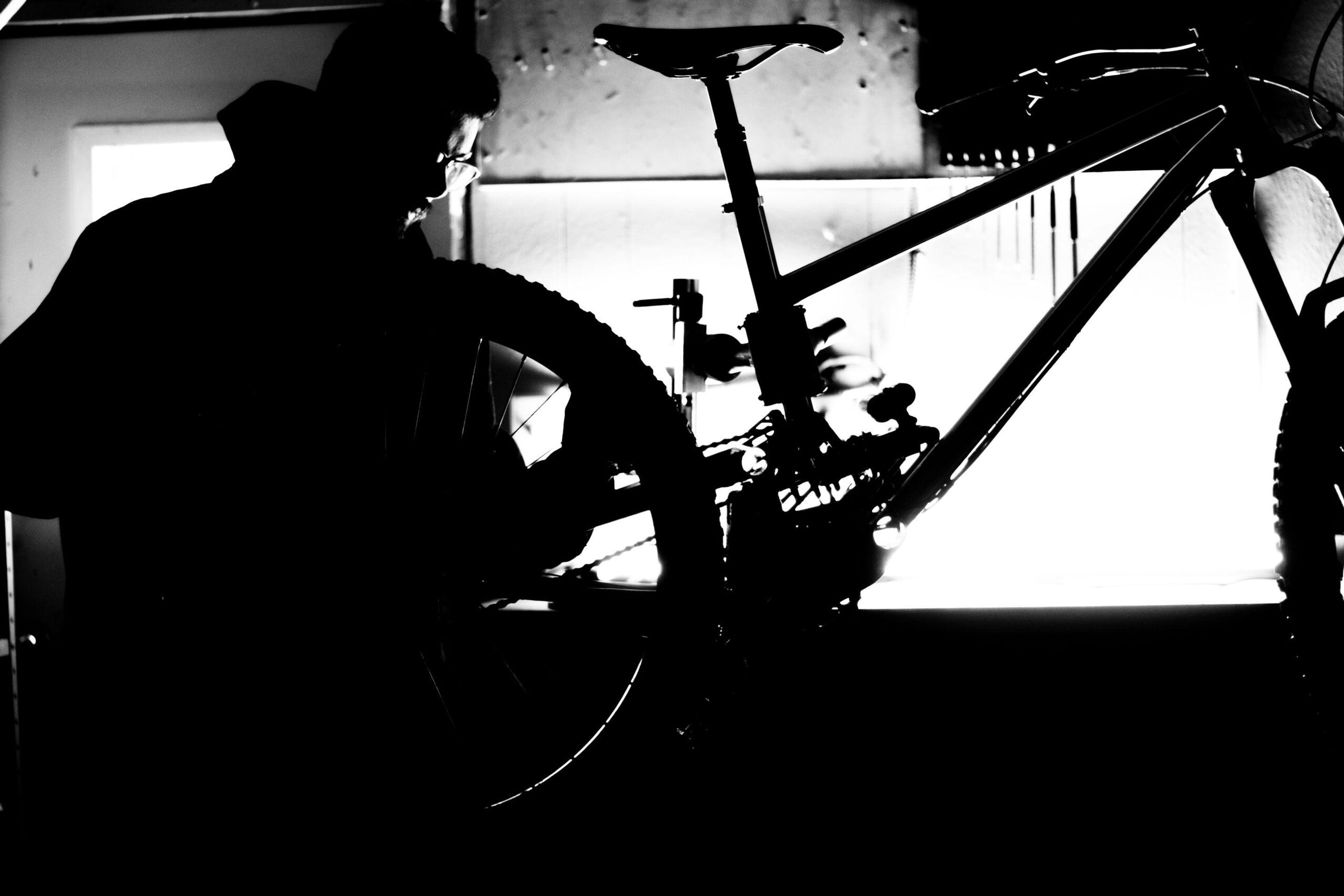 Black and white silhouette of a craftsperson assembling or welding a mountain bike frame in a studio workshop, backlit by bright window light.