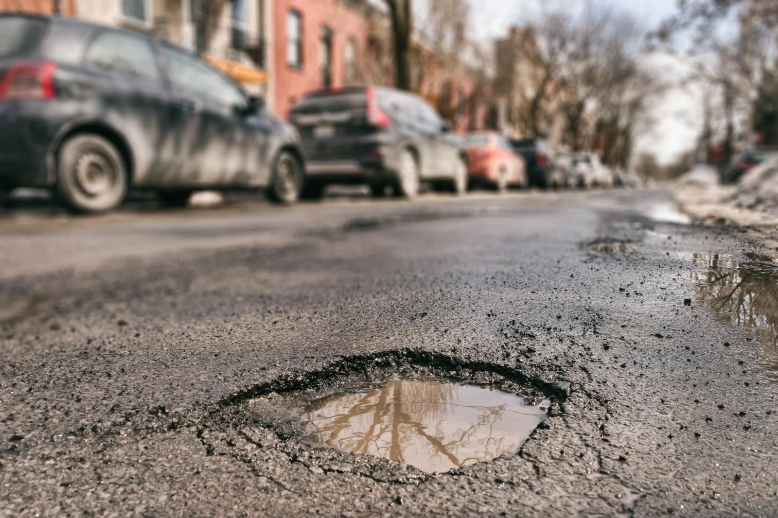 Close-up of a large pothole filled with water on an urban street, with parked cars and buildings visible in the background.