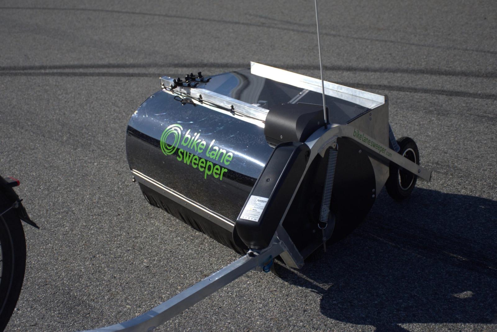 Close-up of an electric bike lane sweeper attachment showing the active brush mechanism, motor mount, and hopper system designed to clear debris from cycling lanes.
