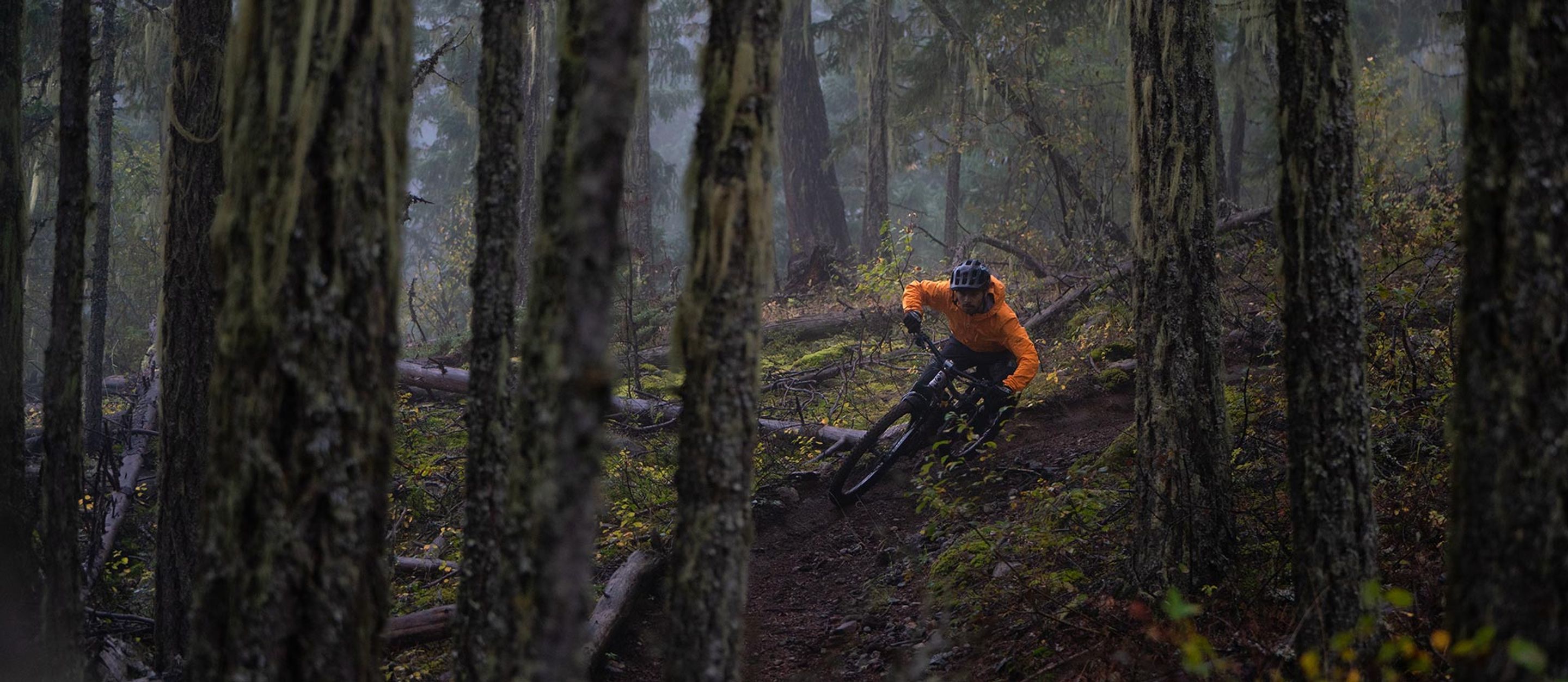 Mountain biker in an orange jacket leaning into a downhill turn on a misty forest trail lined with moss-covered trees.