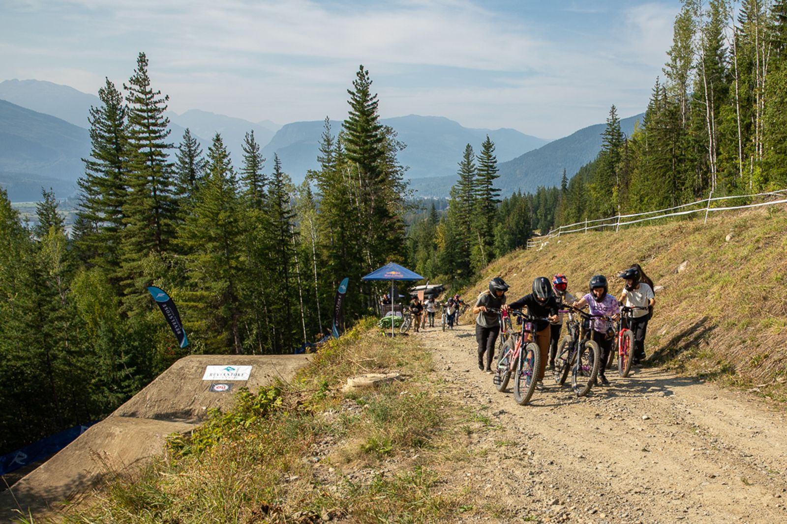 Female riders gathering with their bikes at the Dark Horse freeride event at Revelstoke Mountain Resort, with jump features, spectator tent, and snow-capped mountains in the background.