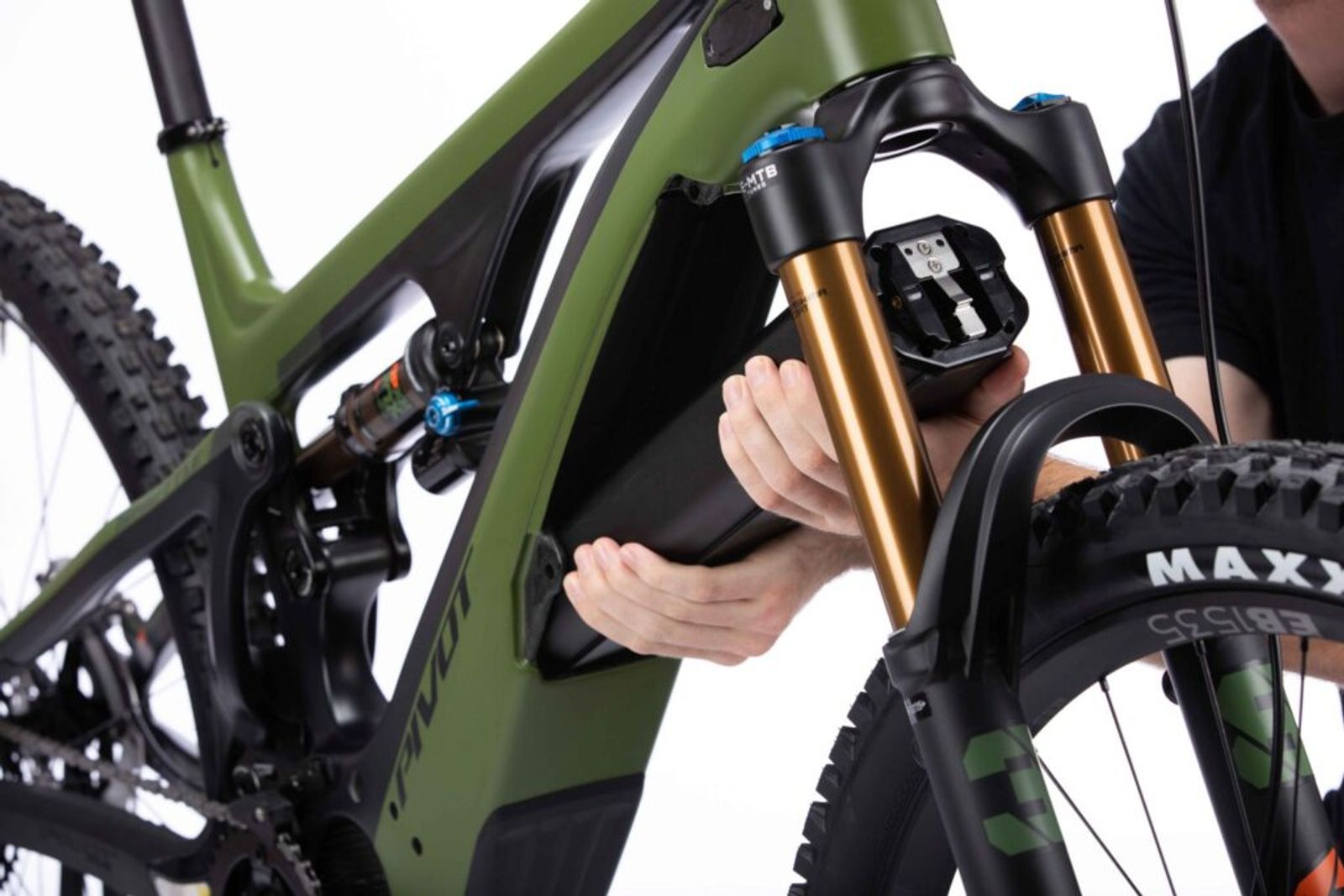 Close-up of a person's hands removing or installing an electric bike battery on a green mountain bike with bronze suspension forks against a white background.