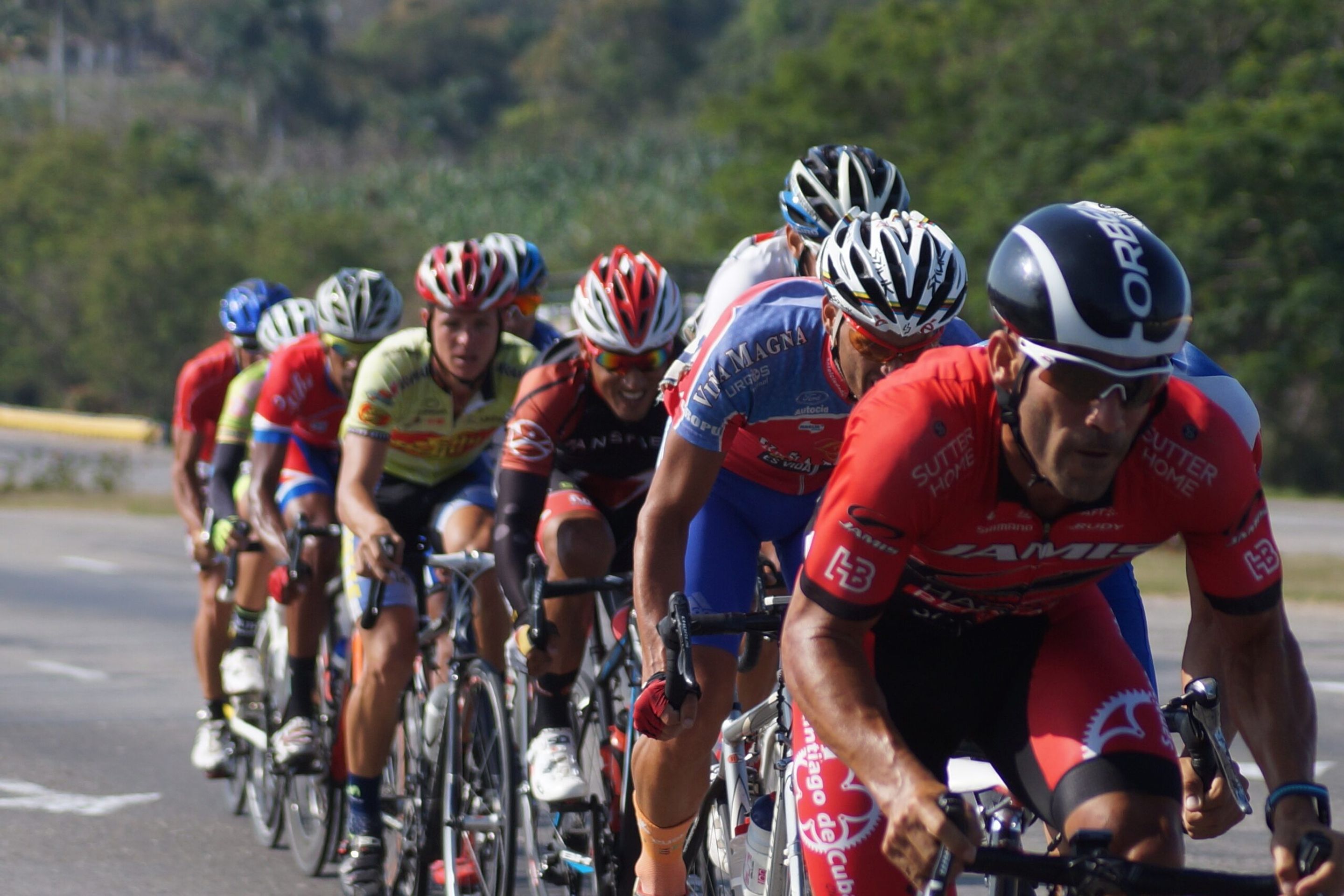 Group of cyclists riding together during a Gran Fondo event, dressed in team cycling kits with helmets, riding in formation on a road with trees in the background.