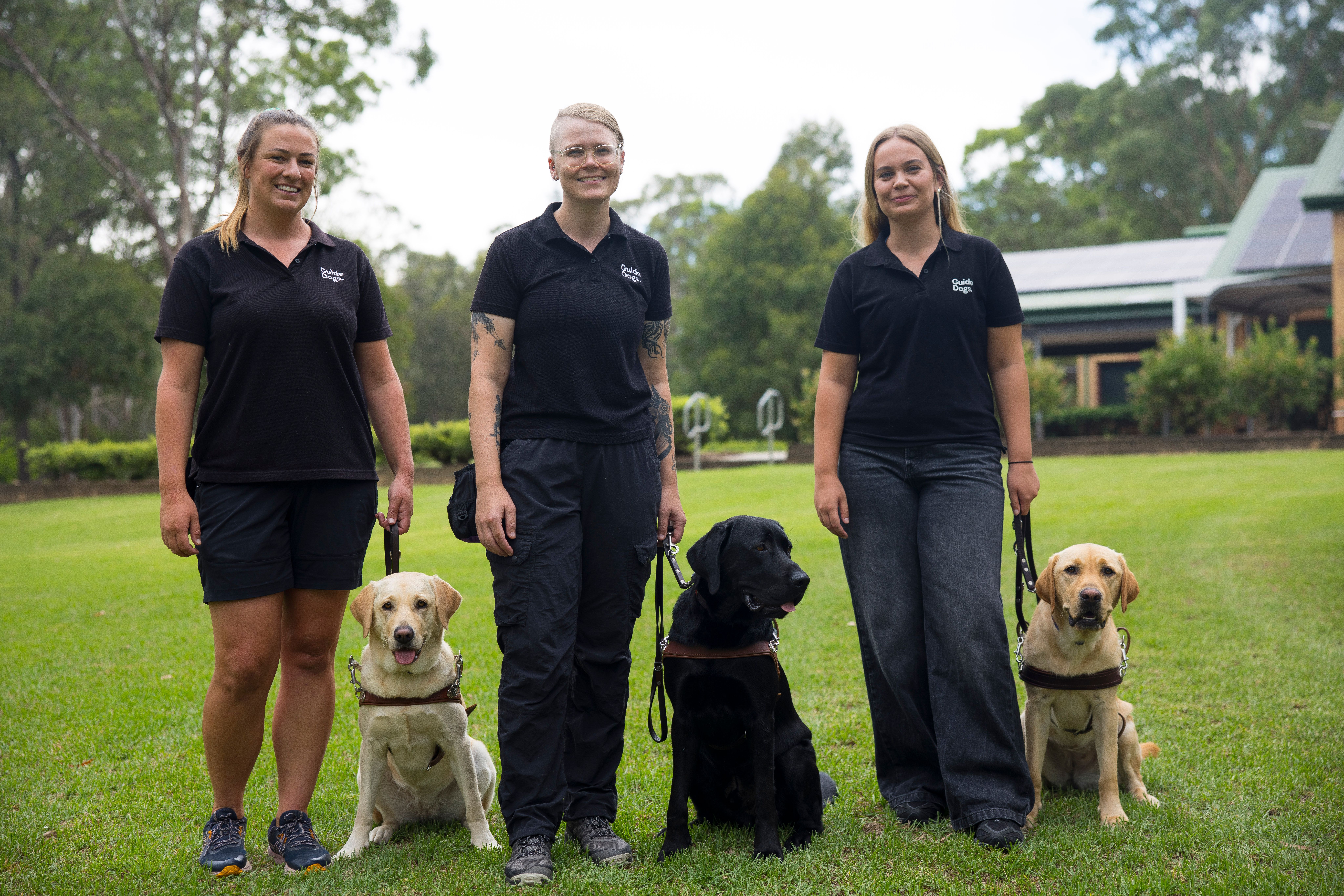 Three Guide Dog Trainers standing each with a Labrador sitting next to them.