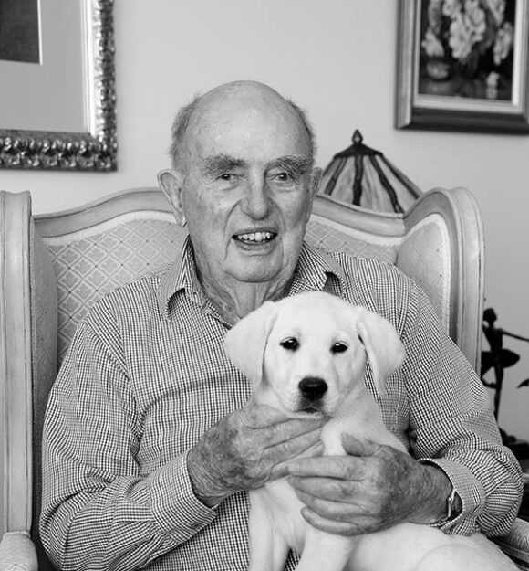 Black and white image of Doug Cameron holding a yellow Labrador puppy on his lap.