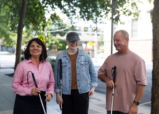 Three Clients stand together smiling and holding their White Canes in their right arms.