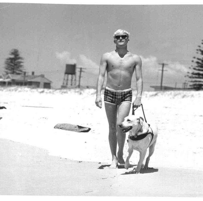 A black and white image of a man walking on the beach with a yellow Labrador Guide Dog.
