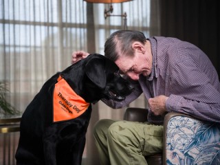 A man resting his head against a black Labrador Therapy Dog.