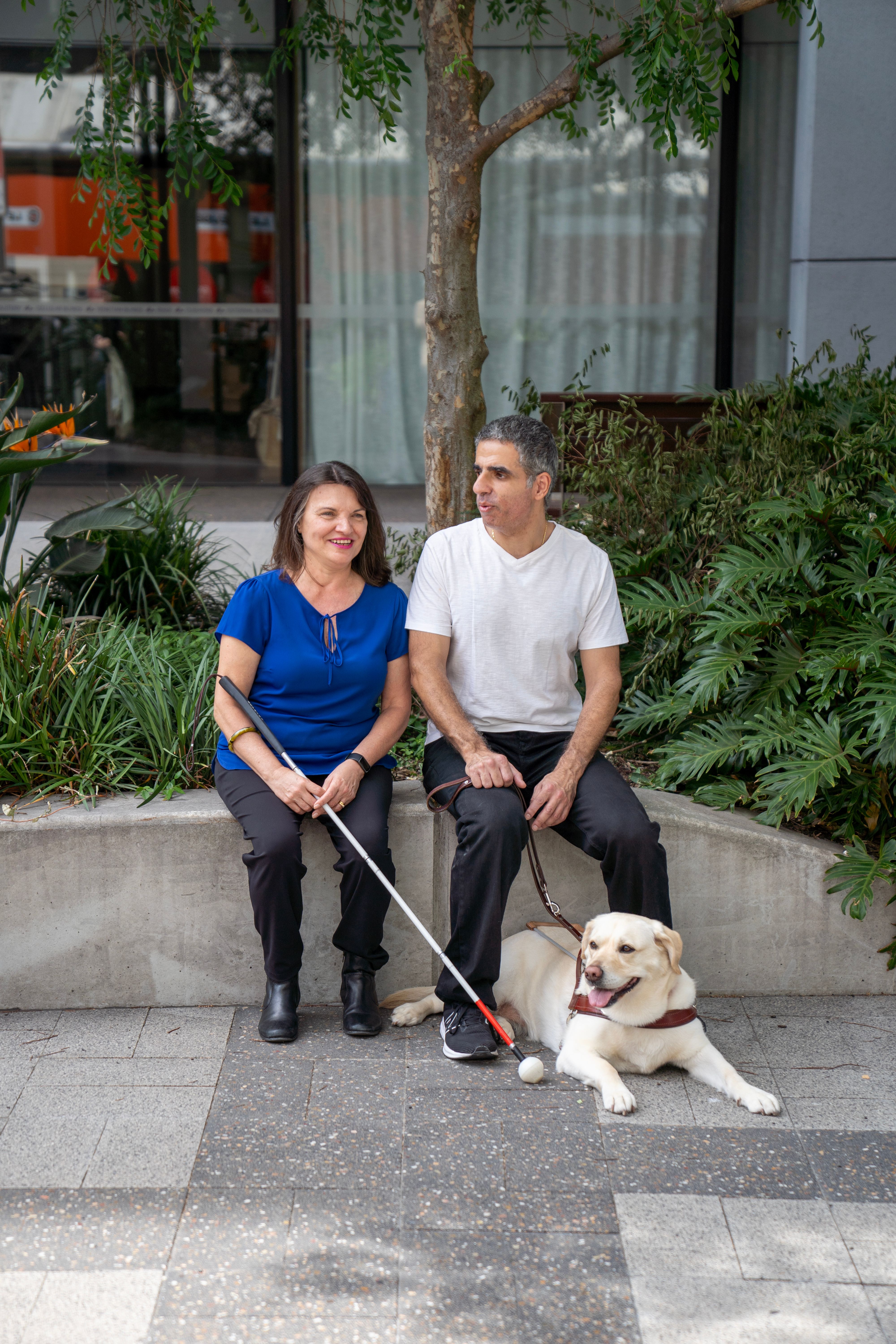 Two people one with a cane and one person with a Guide Dog sitting together.