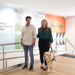 A person guiding a Guide Dog Handler and their Guide Dog.