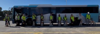 Eleven people wearing high-vis vests stand outside in front of a bus. There is a Labrador in harness sitting with each person.