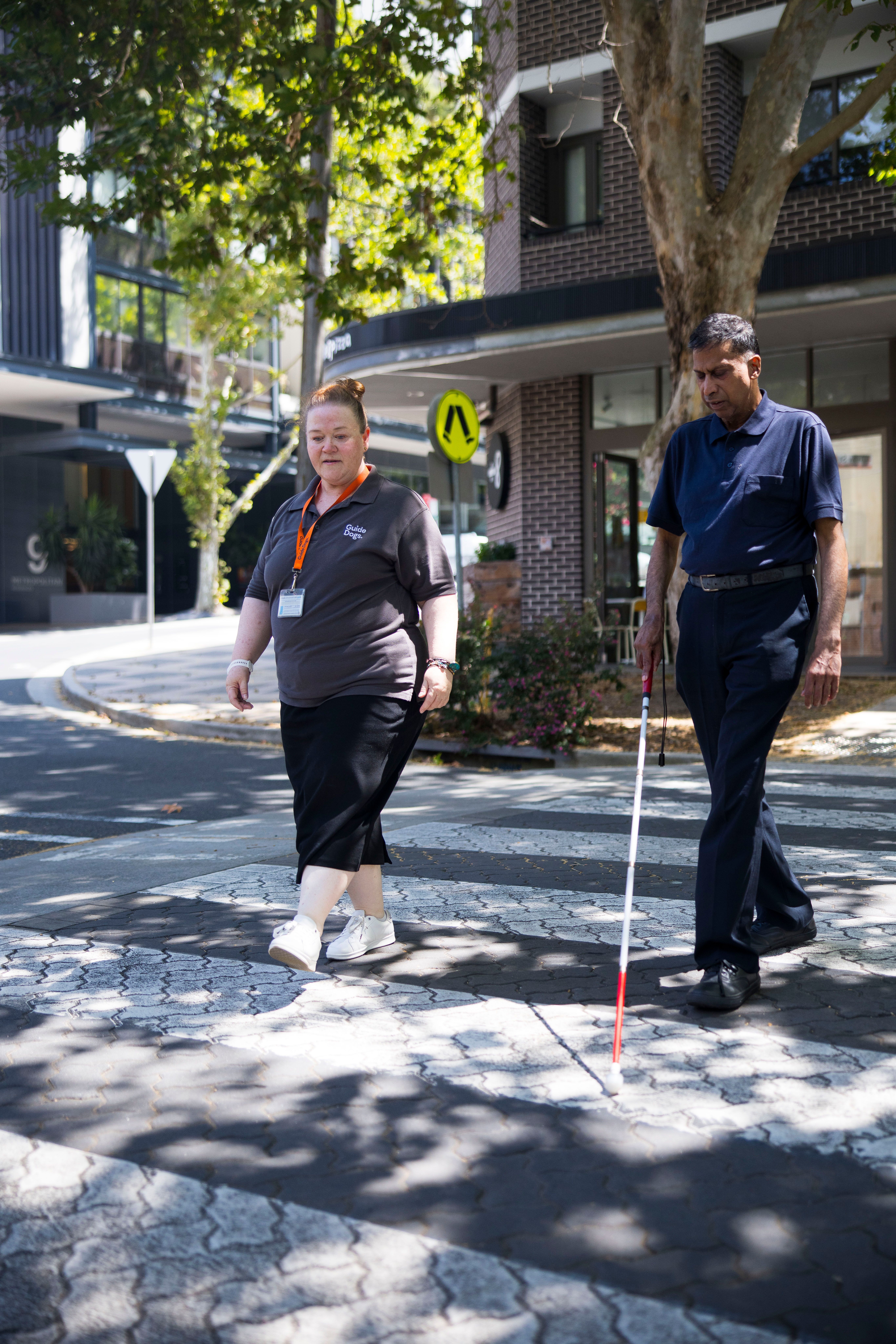 A staff member and Client walking across a crossing with their white cane.