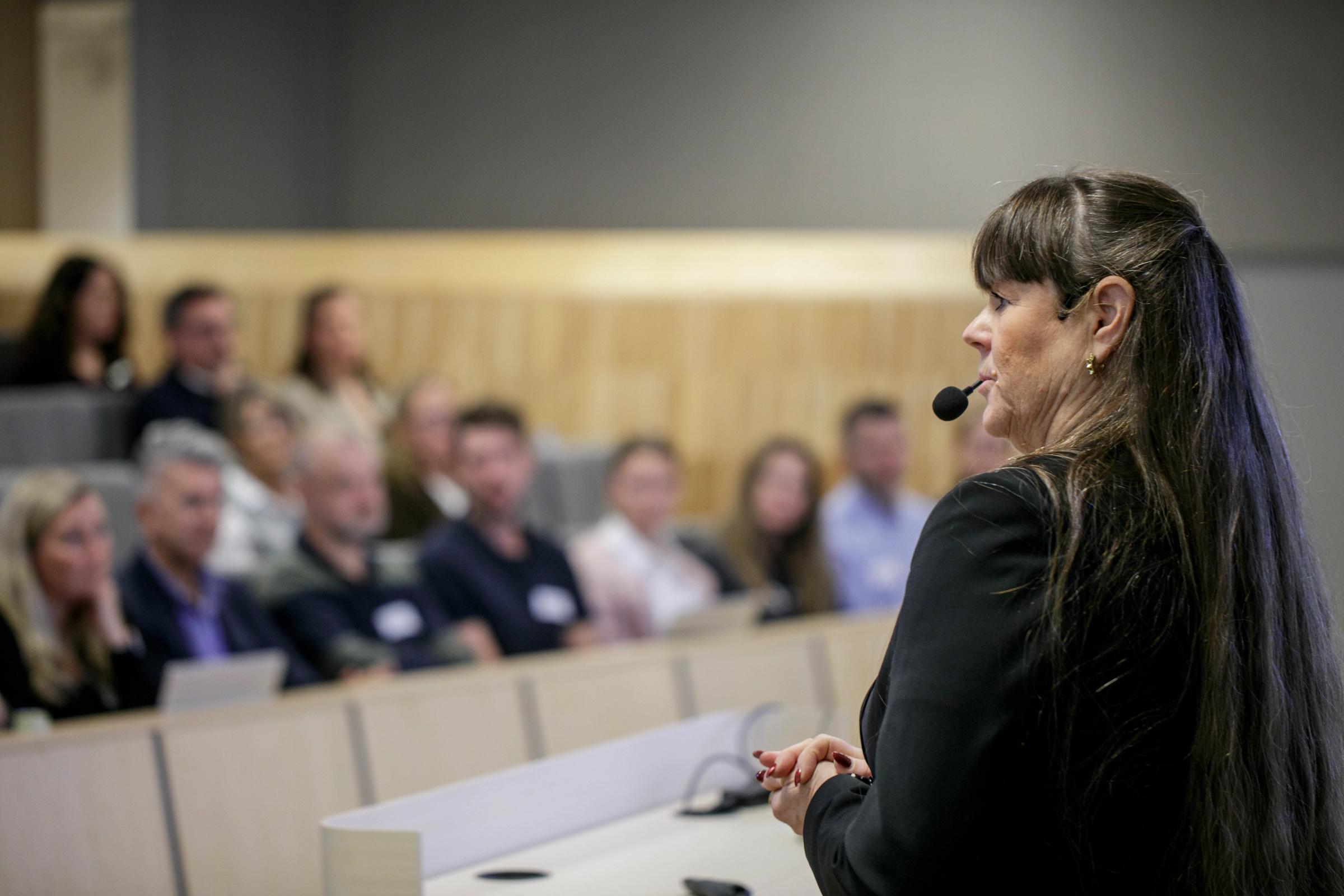En föreläsare som syns i förgrunden med åhörare som sitter i en föreläsningssal