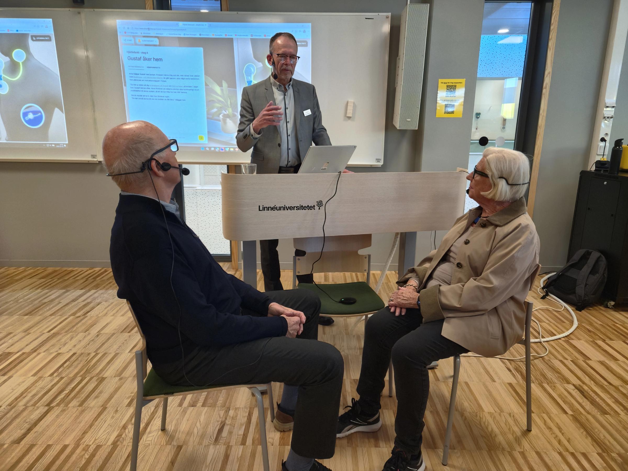 Två personer sitter mittemot varandra vid ett litet bord och lyssnar på den tredje personen i bilden som ger instruktioner