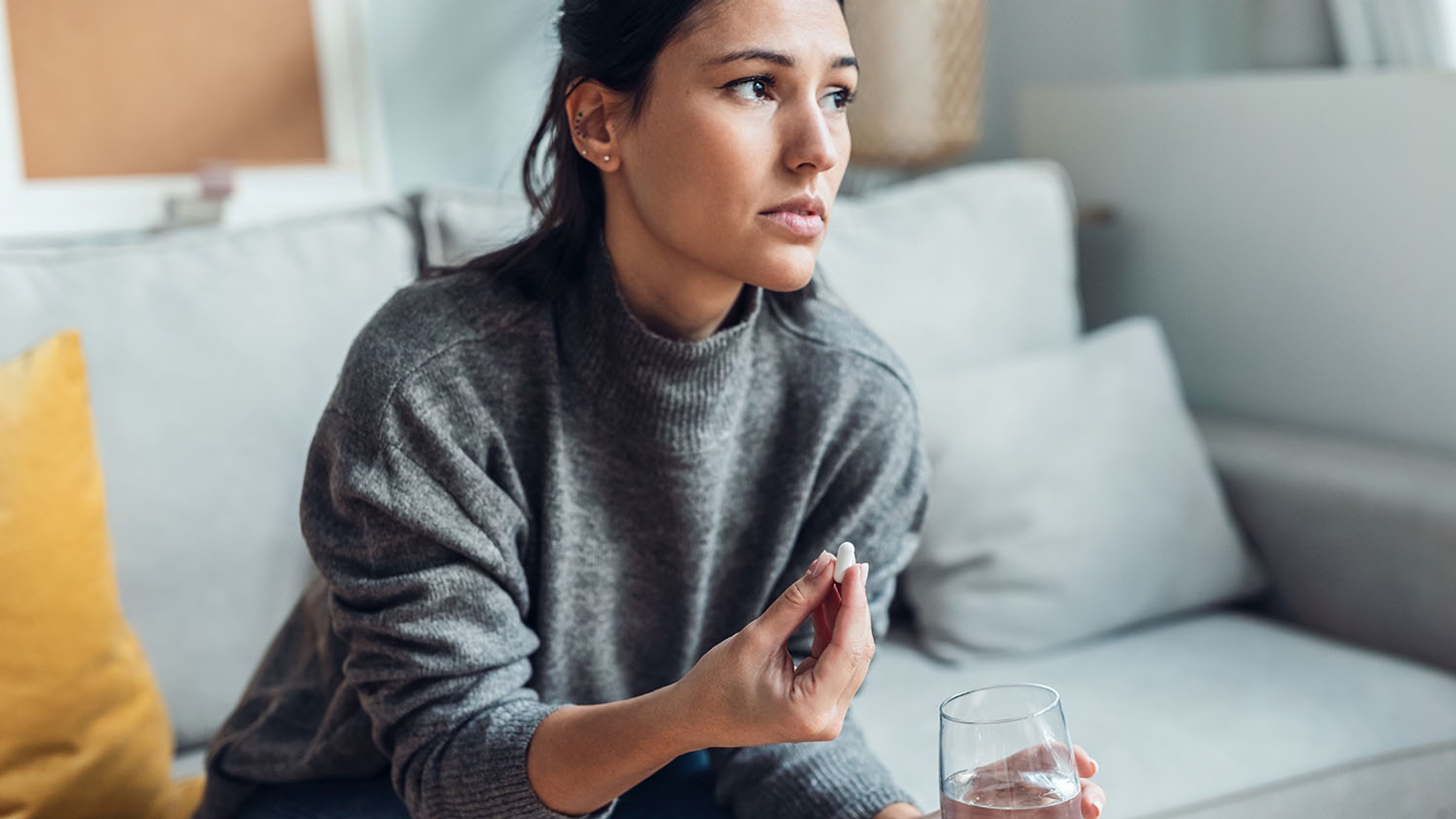 Woman with a pensive expression holding a pill and a glass of water.