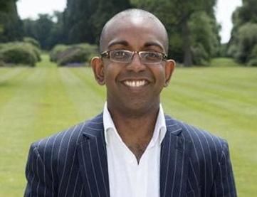 Smiling bald man wearing glasses and a pinstripe suit standing in a park.