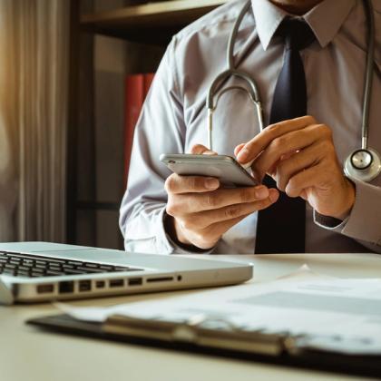 Doctor in a white coat and tie using a smartphone at a desk with a laptop and stethoscope in the background