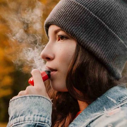 Young woman in a gray beanie and denim jacket vaping outdoors with autumn leaves in the background