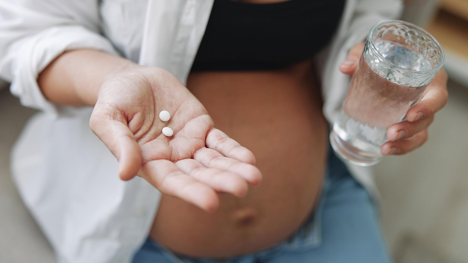 A pregnant person holds two white pills in one hand and a glass of water in the other, with their bare belly visible.