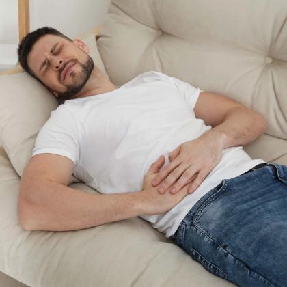Man lying on a couch holding his stomach in pain, looking uncomfortable and grimacing.