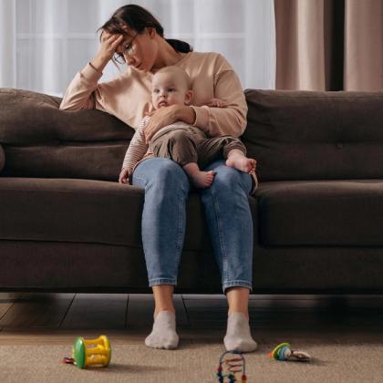 Tired mother sitting on a couch holding a baby, surrounded by scattered toys, expressing exhaustion.