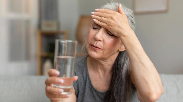 A woman with gray hair holds a glass of water and presses her hand to her forehead, appearing unwell.