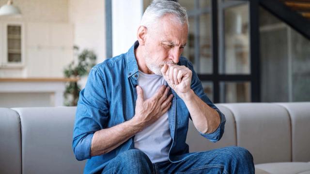 Elderly man coughing into his fist while holding his chest.