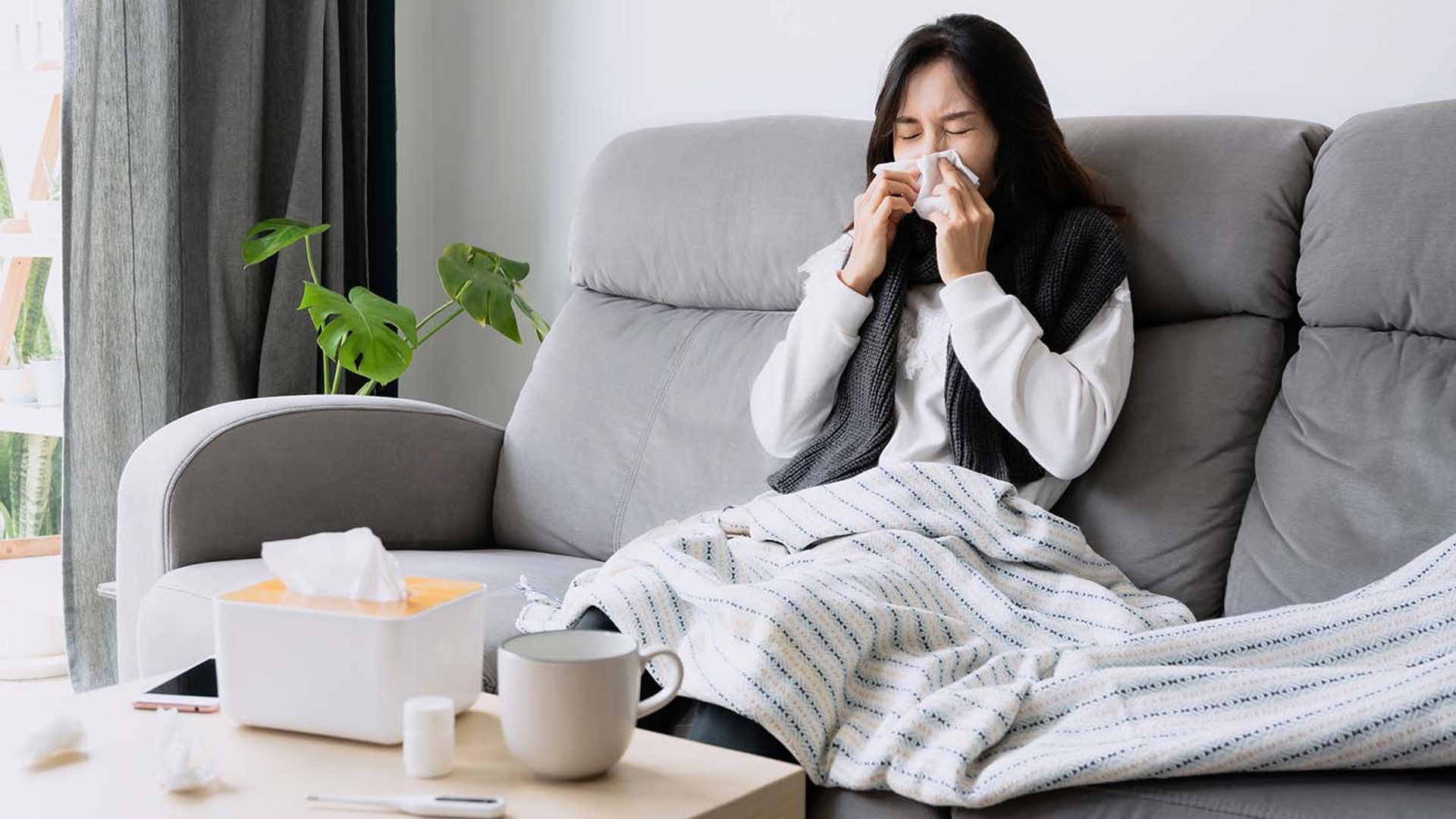 A woman wrapped in a blanket sits on a sofa, blowing her nose with a tissue.