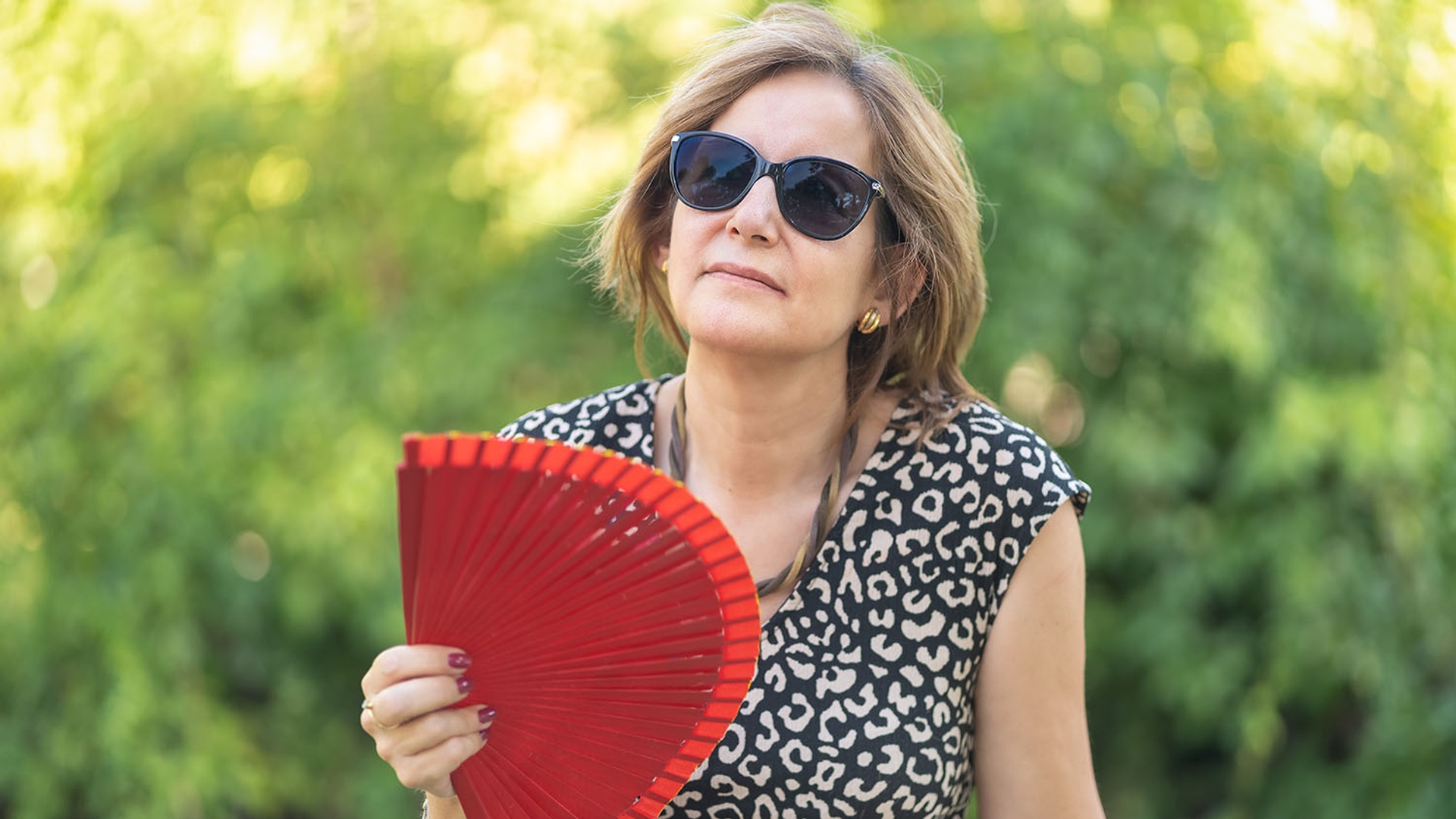 Woman wearing sunglasses and holding a red hand fan outdoors.