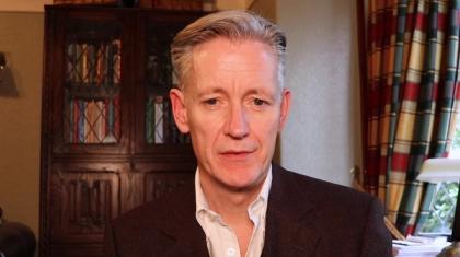 Middle-aged Caucasian man speaking in a traditional room with stained glass cabinet and plaid curtains in the background.