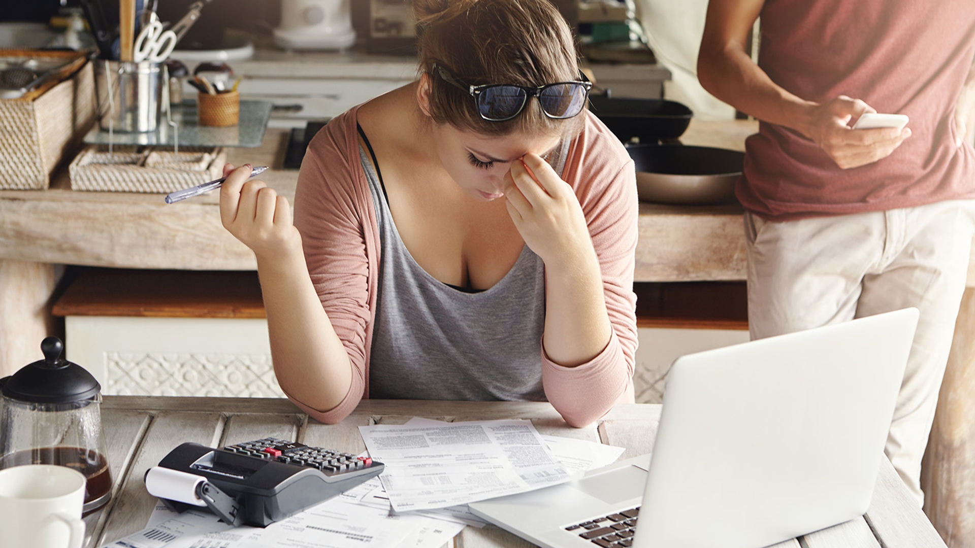 Woman feeling stressed while managing finances with calculator, laptop, and bills on kitchen table, man in background using smartphone