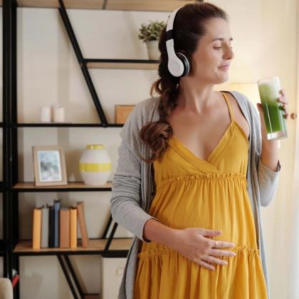 Pregnant woman in a yellow dress and headphones smiling and holding a green smoothie in a modern living room