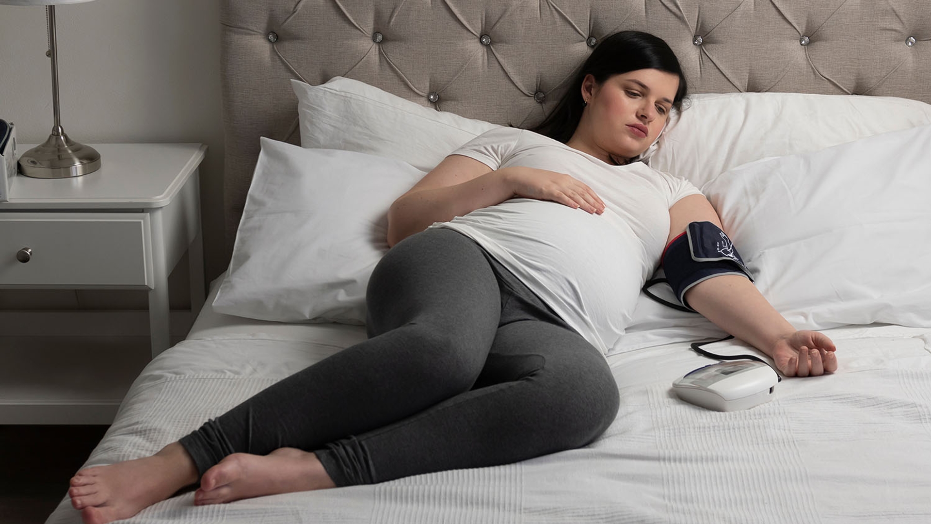 A pregnant woman lies in bed, checking her blood pressure with a monitor.