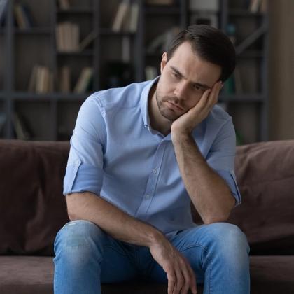 Young man looking thoughtful while sitting on a couch with his cheek resting on his hand, in a living room with bookshelves in the background