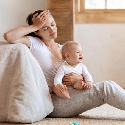 Worried mother sitting on the floor holding her crying baby with a hand on her forehead in a soothing home environment