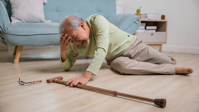 An elderly man lies on the floor, holding his head in distress, after a fall. His cane and glasses are nearby.
