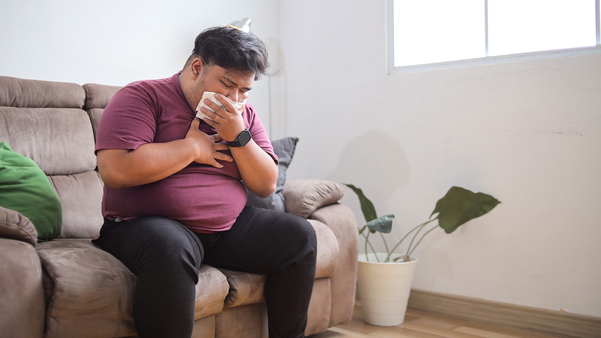 Man on a couch blowing his nose with a tissue and clutching his chest.