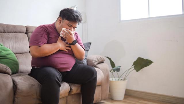 Man on a couch blowing his nose with a tissue and clutching his chest.