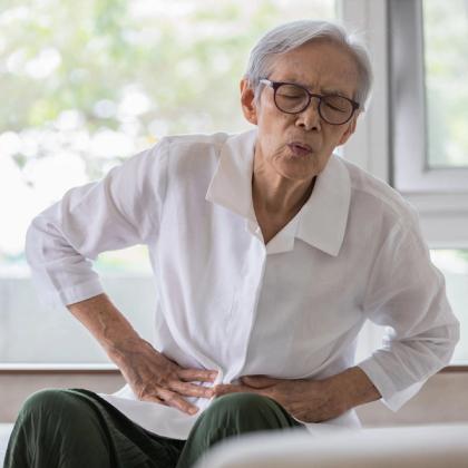 Elderly woman sitting on a couch, touching her side in discomfort or pain, eyes closed and expression showing distress.