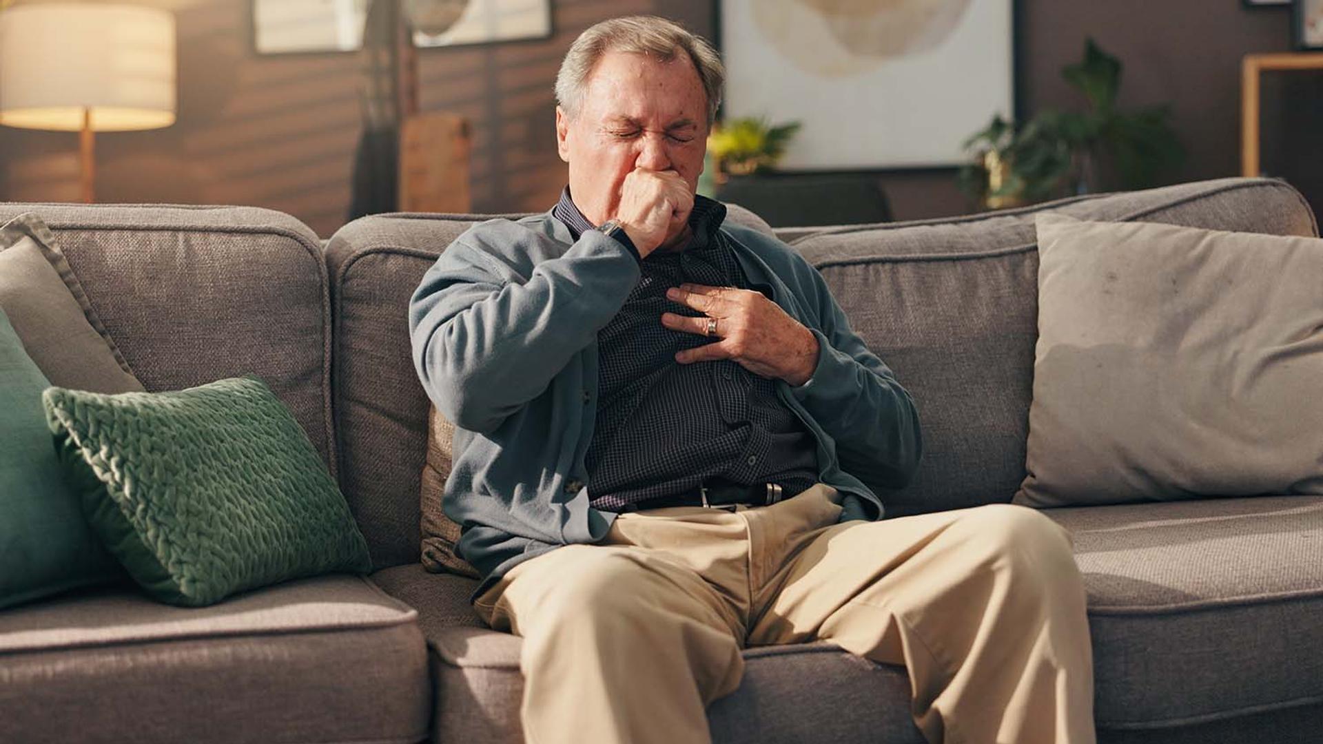 An older man sits on a couch, coughing into his hand and holding his chest.
