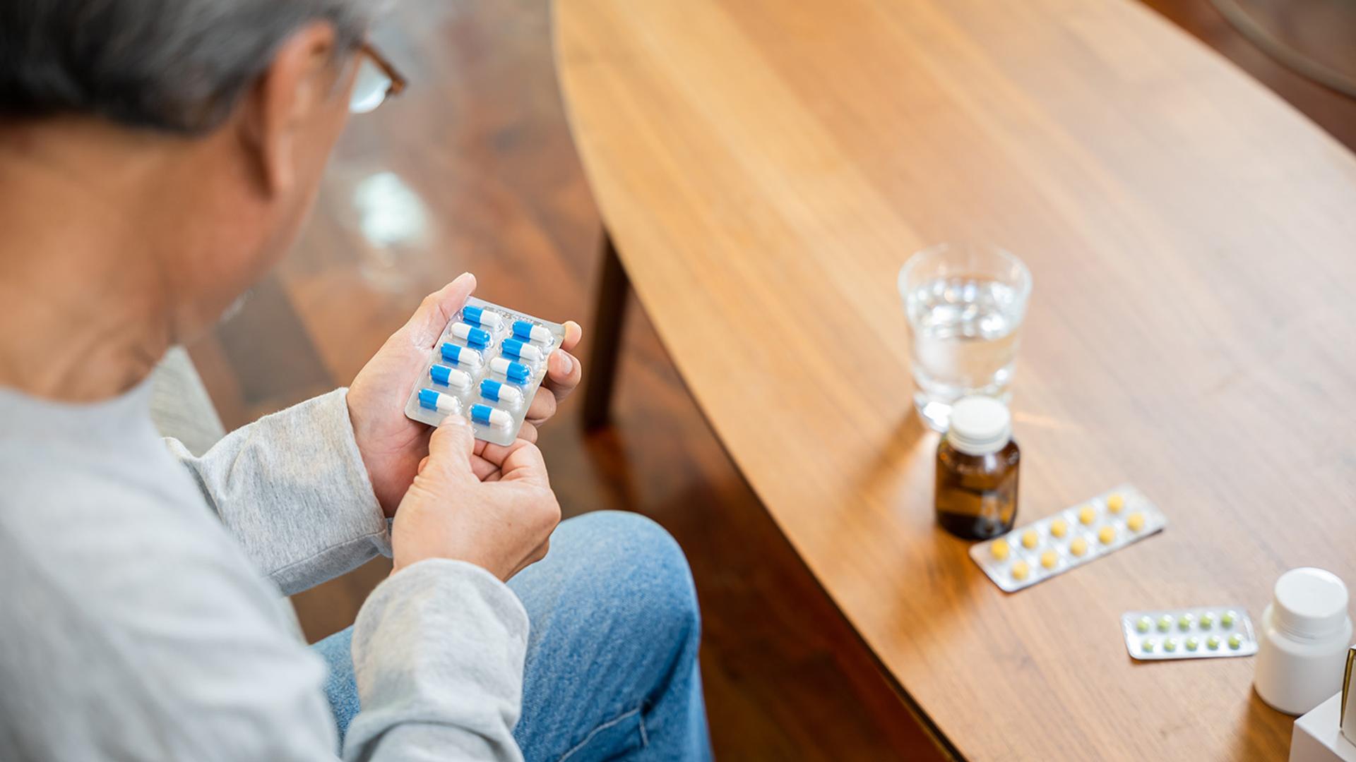 A person with grey hair holds a blister pack of blue and white pills, next to other medications and a glass of water on a table.