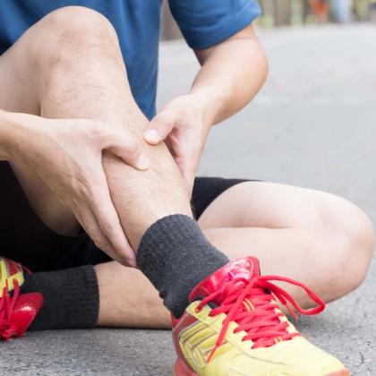 Man sitting on pavement holding his knee in pain, wearing a blue shirt, black shorts, red sports shoes, and a black sock