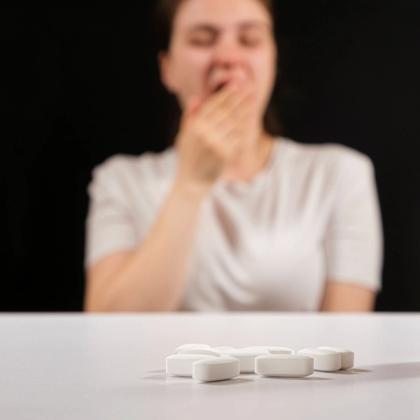 White pills in focus with a blurred person yawning in the background.