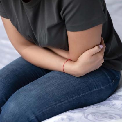 Close-up of a person sitting on a bed, wearing jeans and a dark green t-shirt, hugging their knees with a small red string bracelet on their left wrist.
