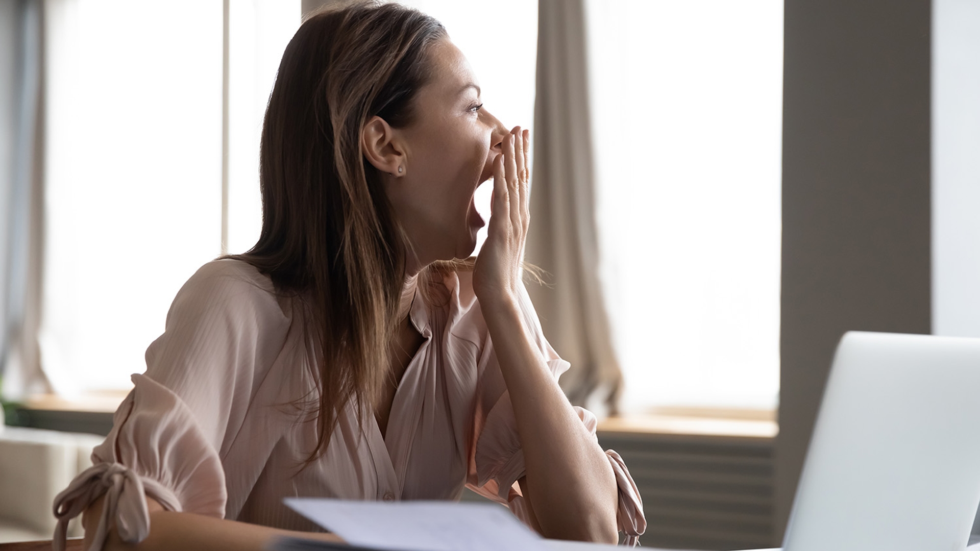 A woman yawns, covering her mouth with her hand, while sitting at a desk.