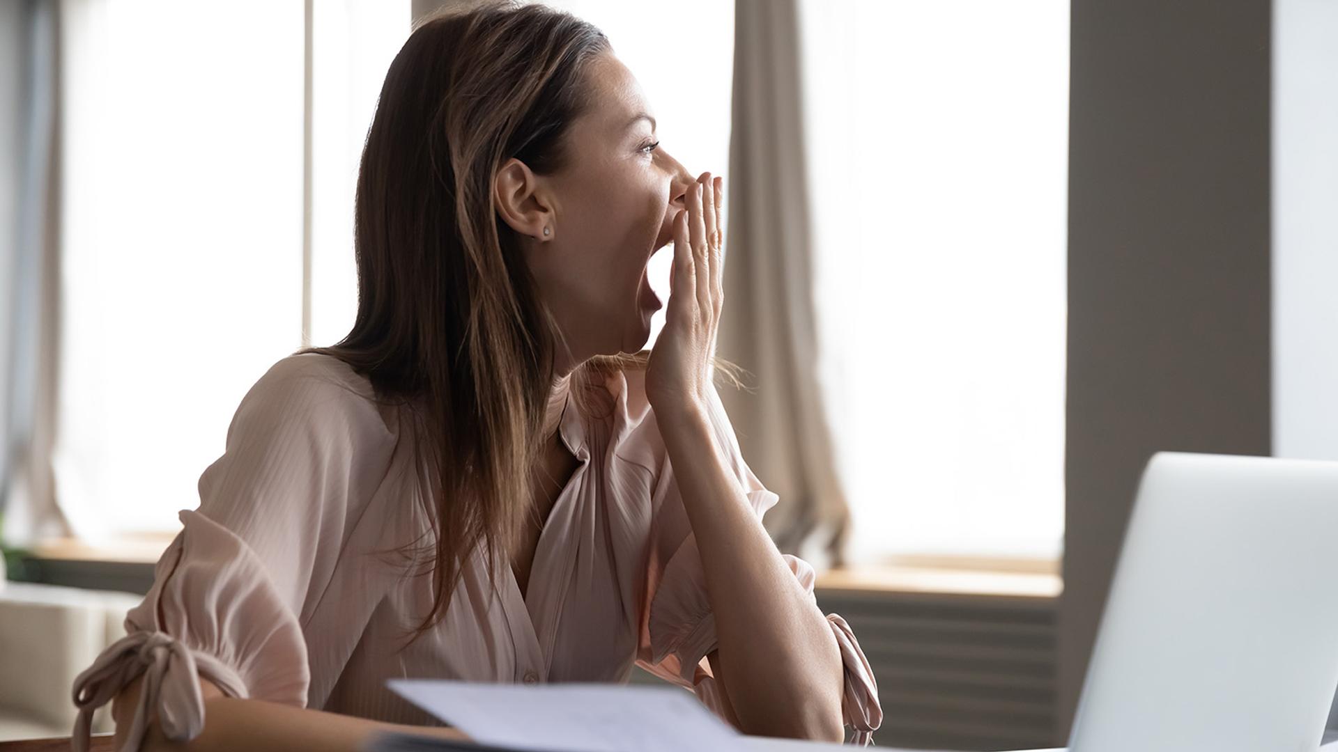 A woman yawns, covering her mouth with her hand, while sitting at a desk.